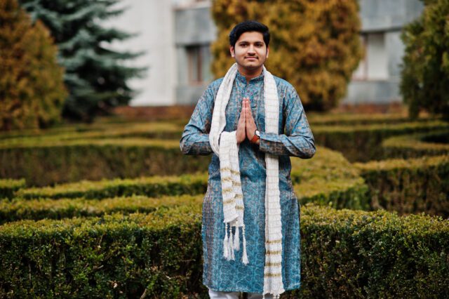 Indian man wear on traditional clothes with white scarf posed outdoor against green bushes at park, show namaste hands sign.