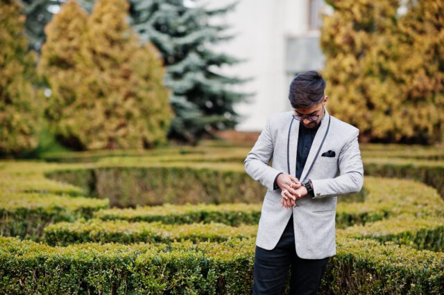 Stylish indian man with bindi on forehead and glasses, wear on suit posed outdoor against green bushes at park and looking at his watches.